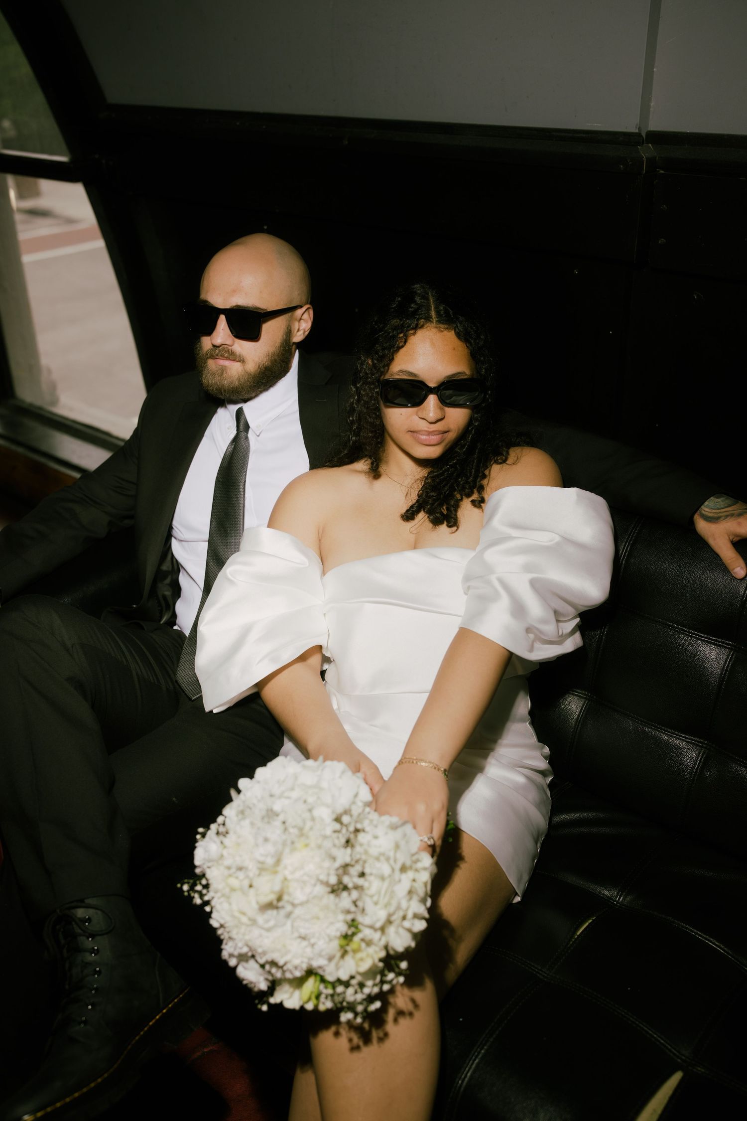 A couple in formal attire sits in a car with white floral bouquet and sunglasses.