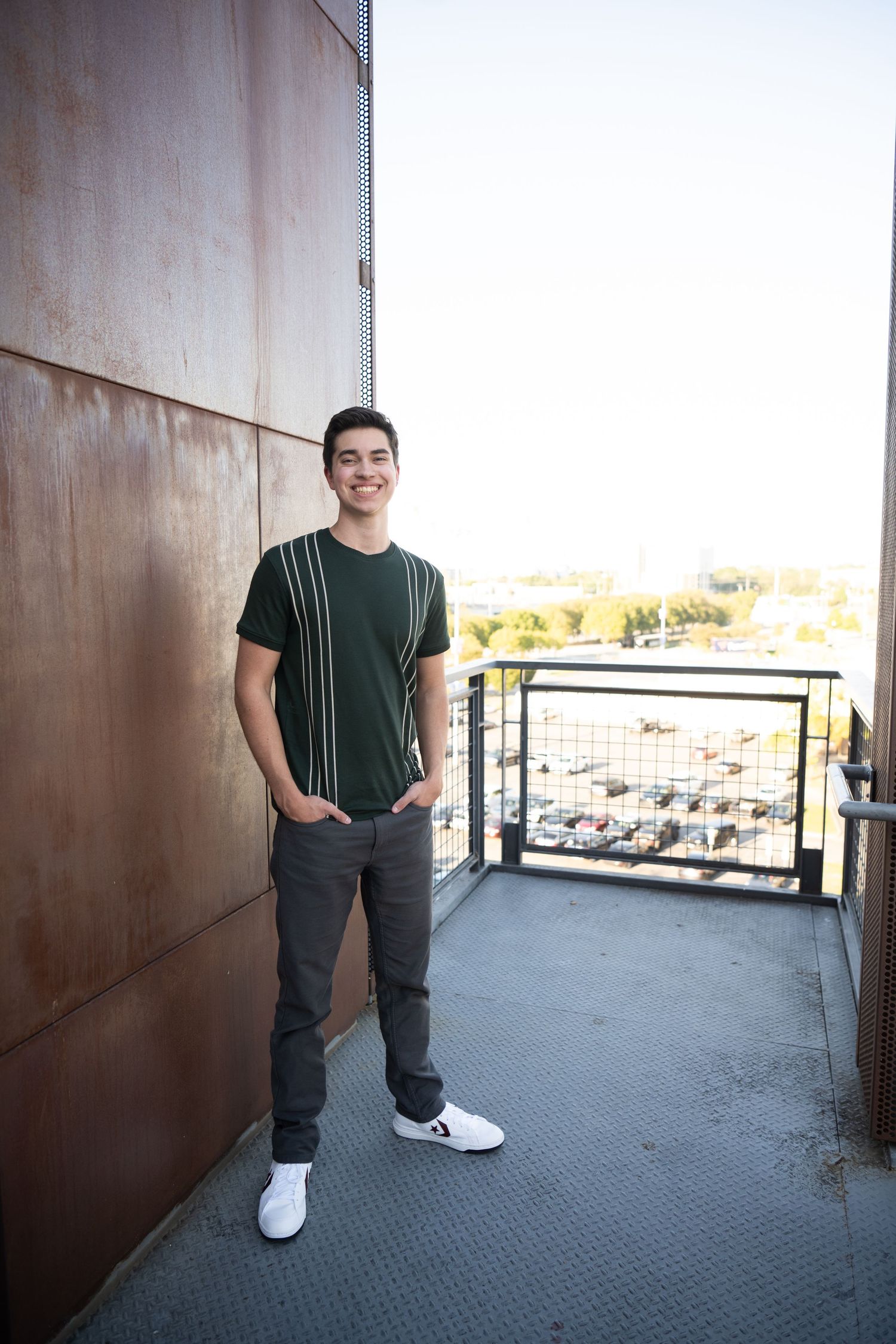 Person wearing dark casual clothing and white sneakers poses in a modern building space with outdoor views.