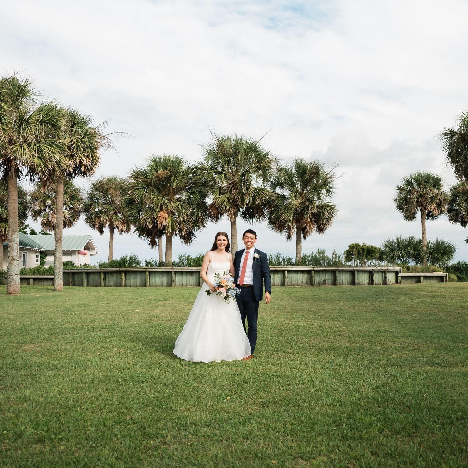 A couple stands on a lush green lawn surrounded by tall palm trees at a scenic outdoor venue.