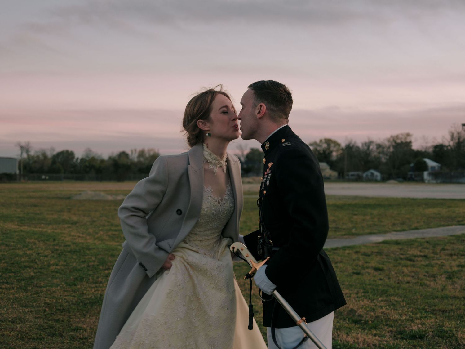A romantic couple in wedding attire shares a kiss in a scenic field at sunset.