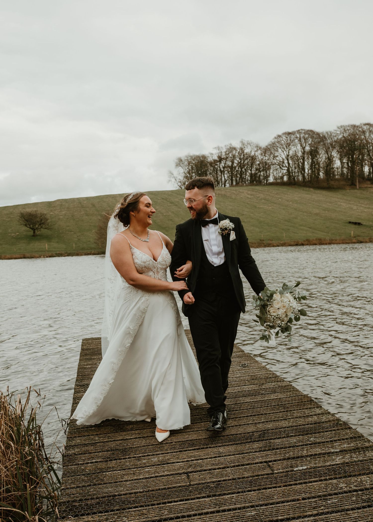 Newlywed couple shares a joyful moment together on a wooden dock overlooking a serene lake on their wedding day.