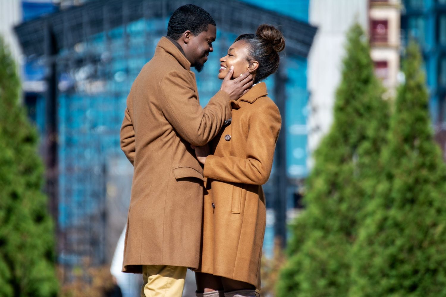A couple in tan coats share a romantic moment against a backdrop of tall buildings and evergreen trees.