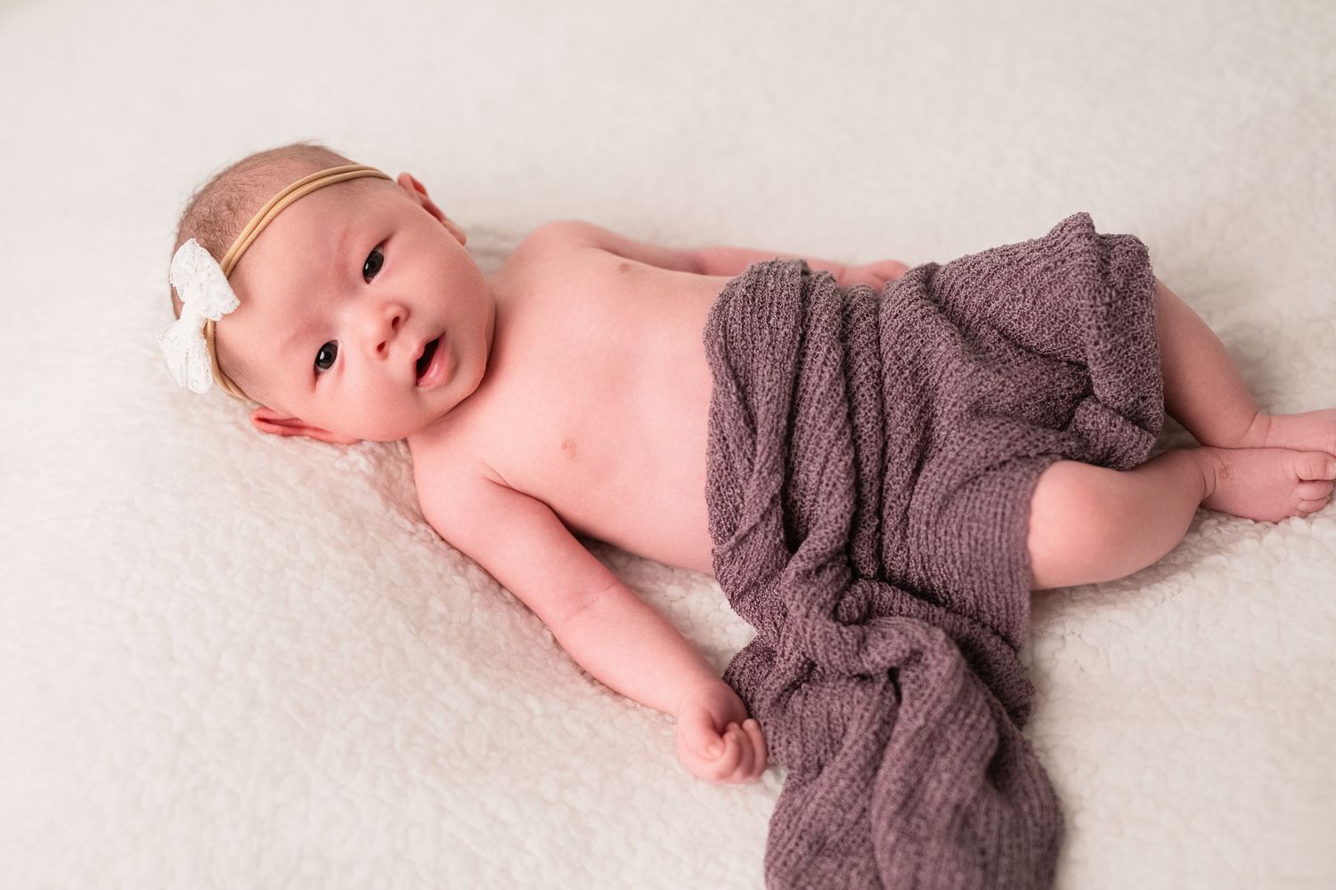 A newborn baby lying on a white blanket while partially wrapped in a soft gray knit blanket.