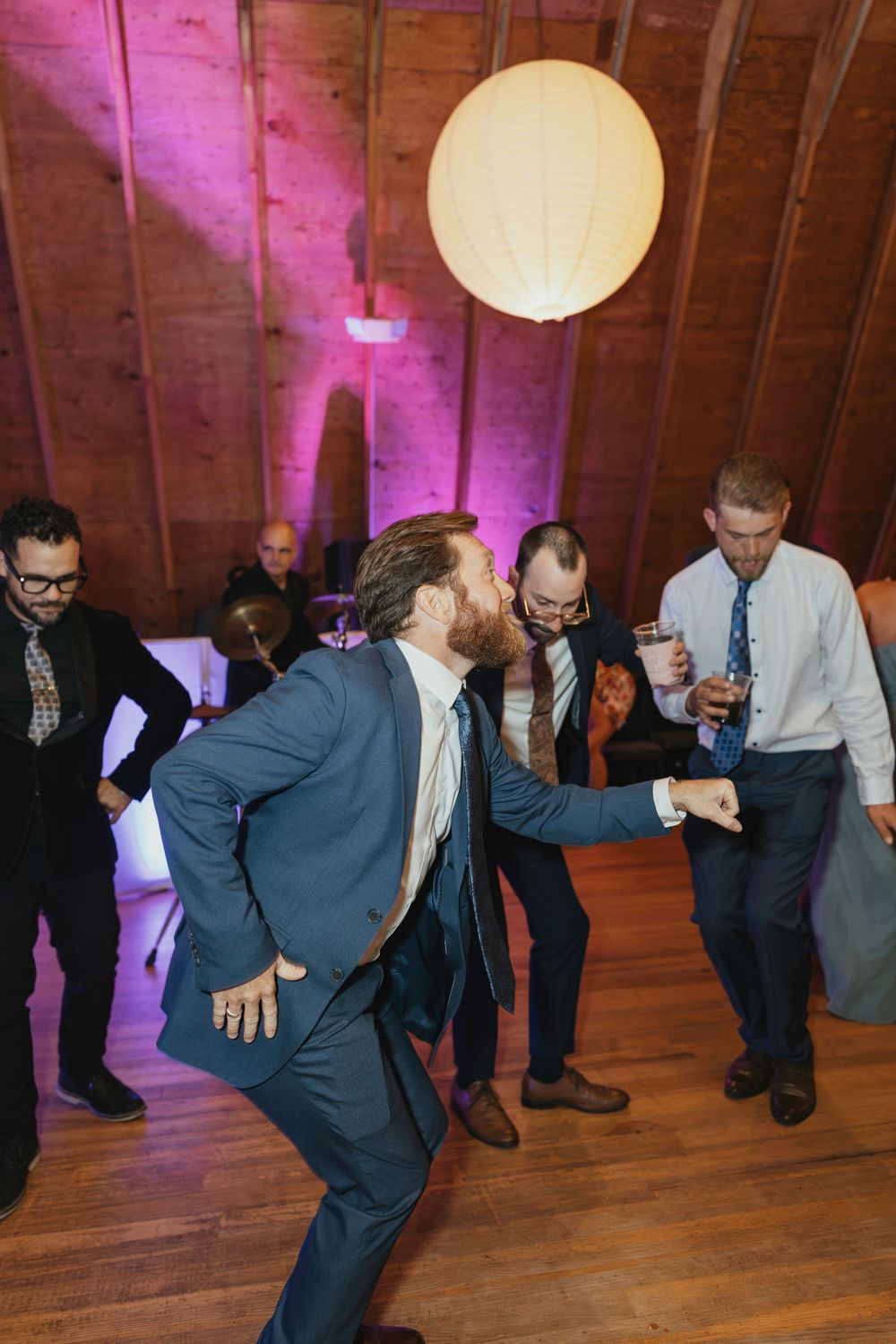 Men in suits perform a synchronized dance move on a wooden floor under paper lanterns with pink lighting.
