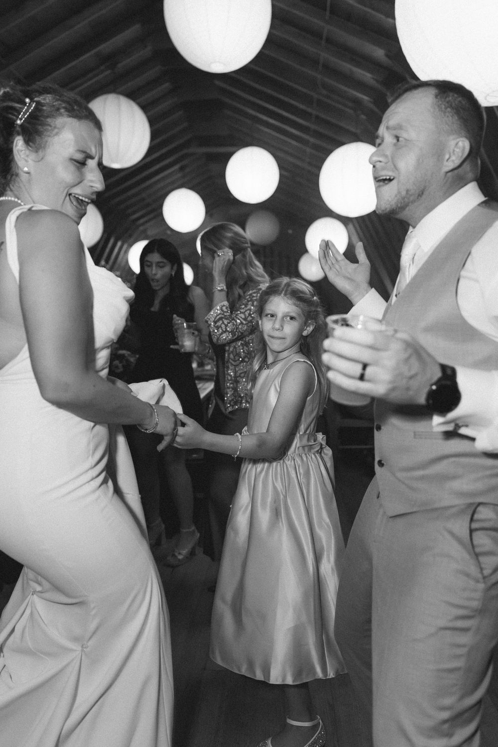 Wedding guests dancing in pairs under white paper lanterns at an indoor reception venue.