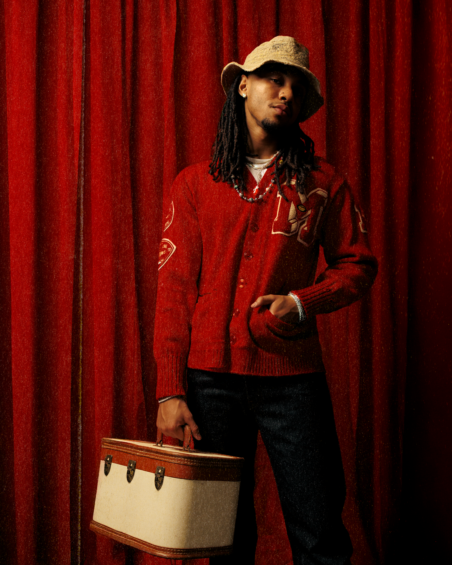 Figure in a red jacket and black pants poses against a red curtain backdrop in a theatrical setting.