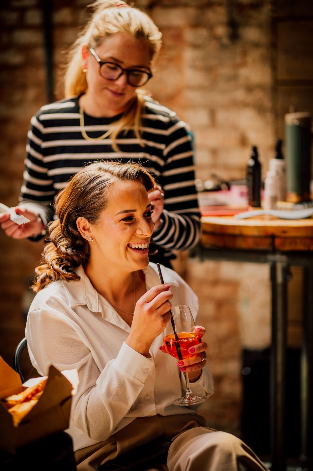 Friends enjoying cocktails and laughing together at a cozy brick-walled bar in warm lighting.