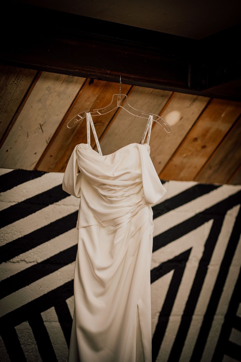 A white wedding dress hangs elegantly against a dark wooden background with dramatic shadows and lighting.