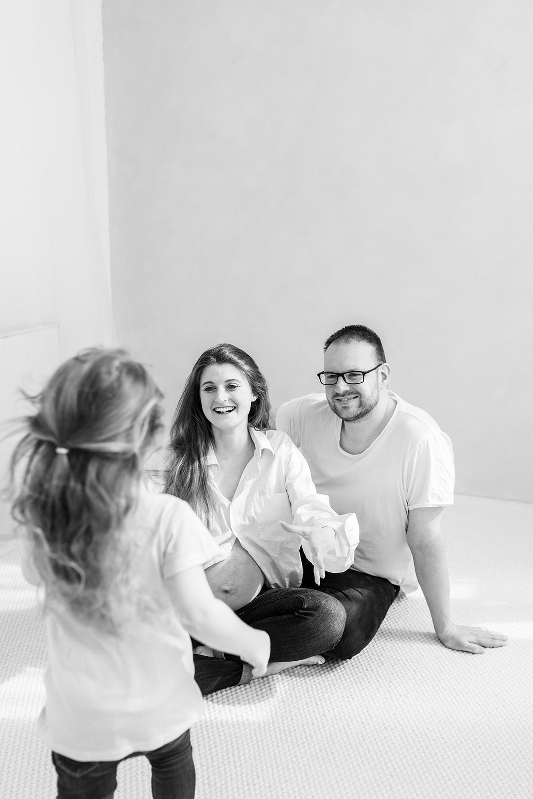 A playful family group sitting and smiling on a white carpeted floor in a bright minimalist space.