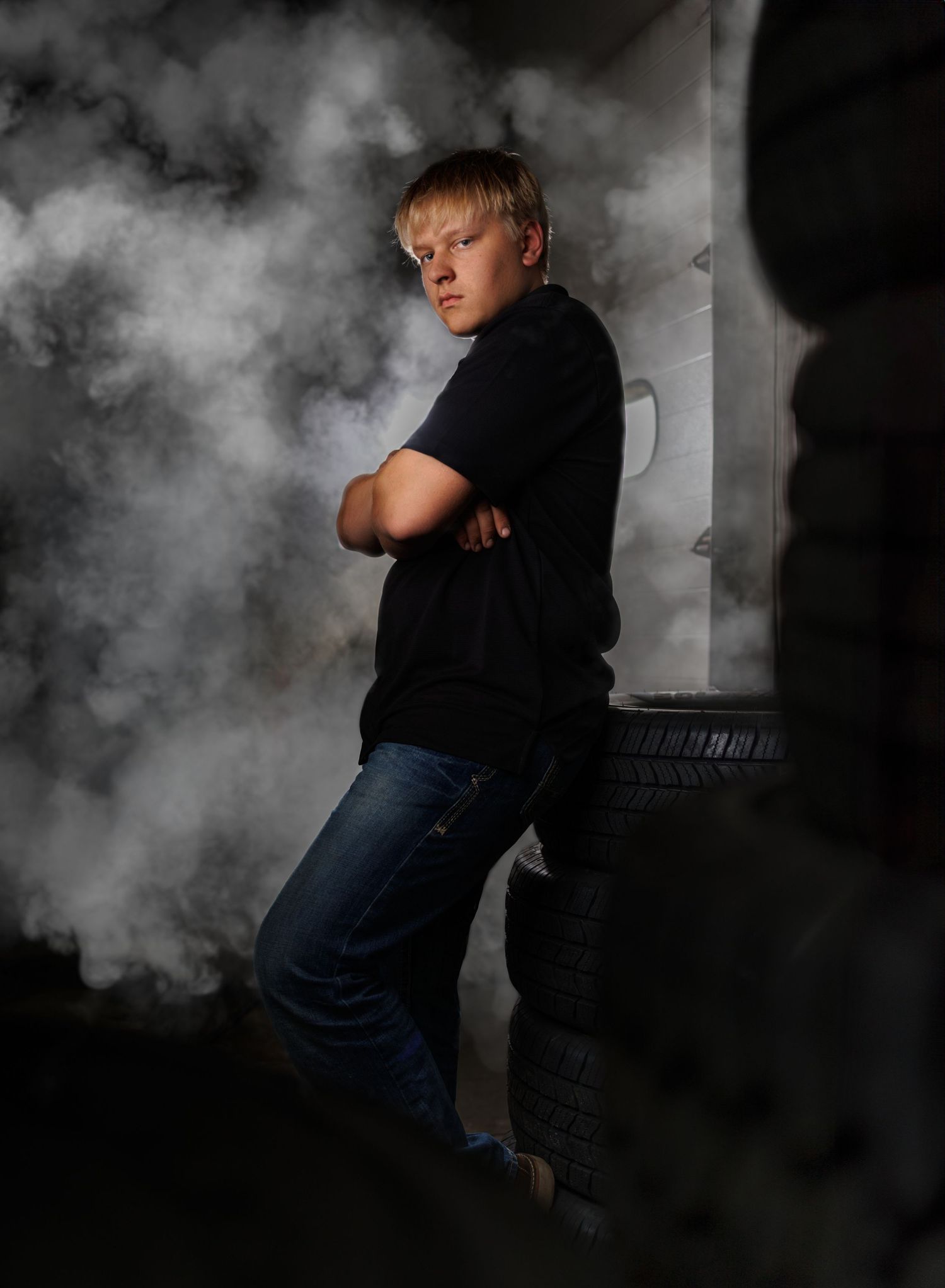 Moody artistic mechanic senior boy portrait with dramatic smoke effects and stack of tires