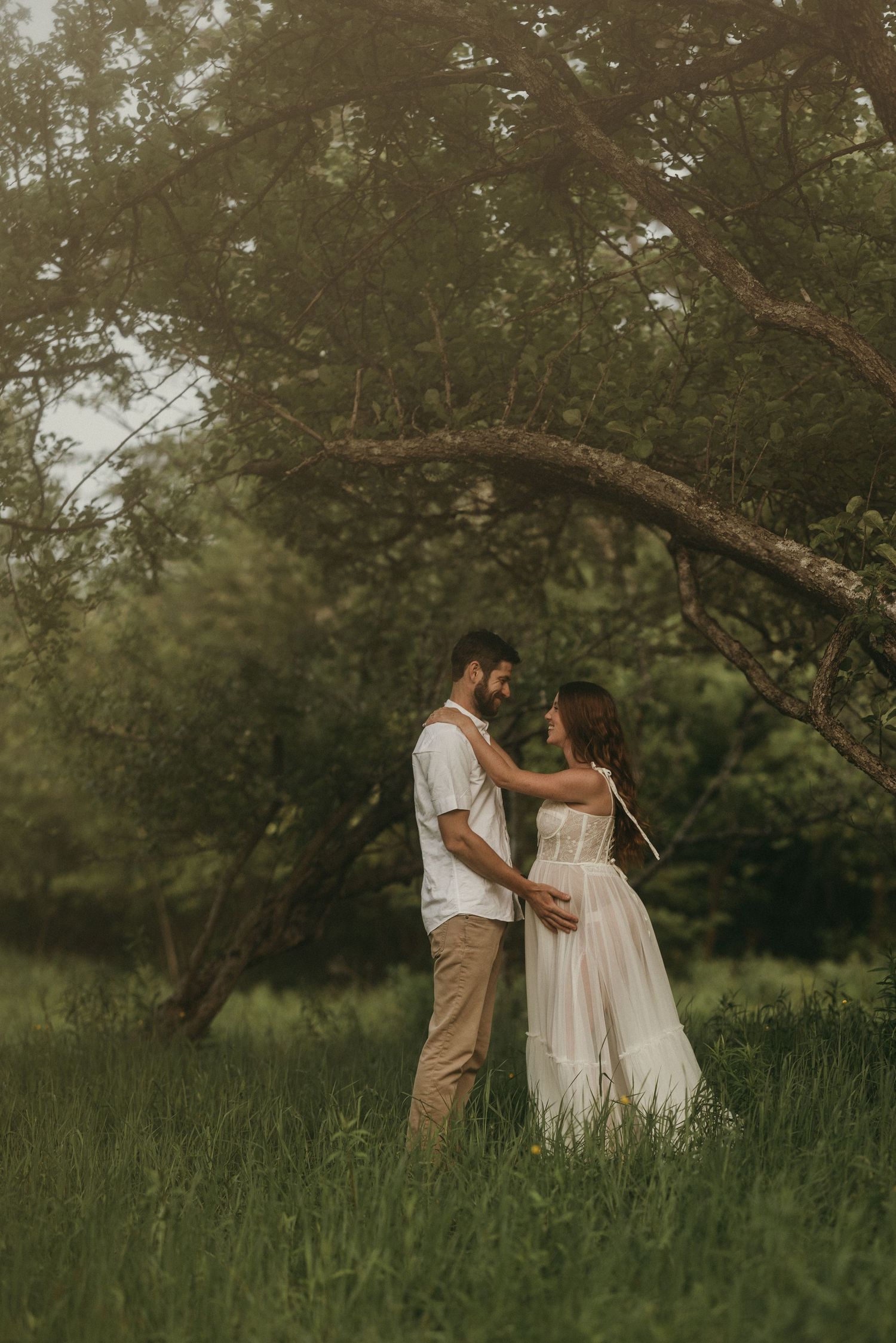 A couple in white dress and khaki attire share an intimate moment in a sunlit meadow surrounded by trees.