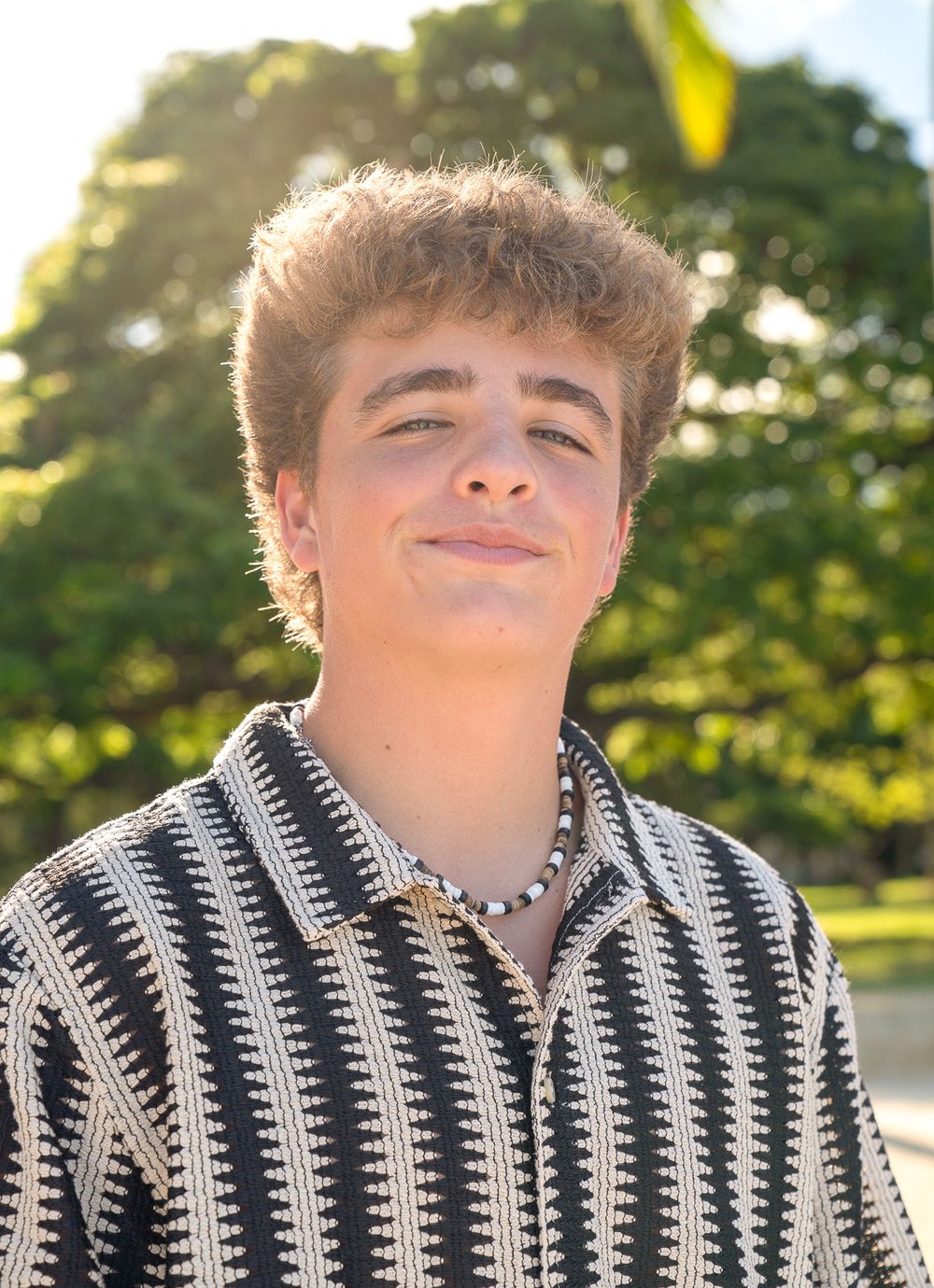 Person wearing black and white striped shirt poses outdoors with sunlit trees in background.