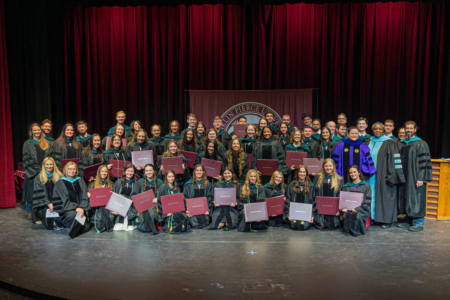 Panoramic view of graduation ceremony with students seated in front of stage with red curtains.