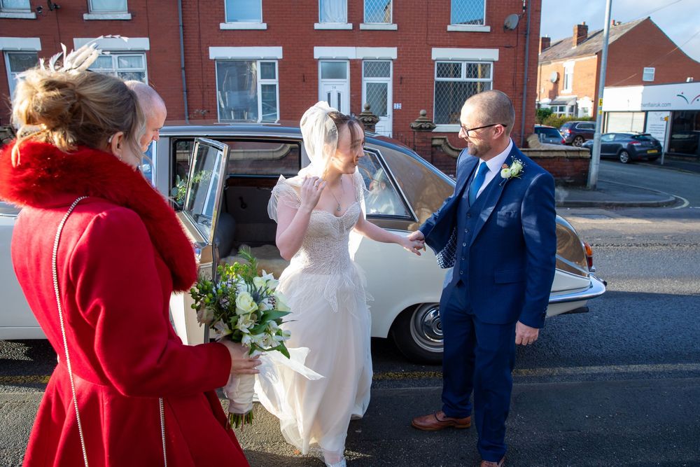 Groom helps bride exit vintage wedding car as guest in red coat watches outside.