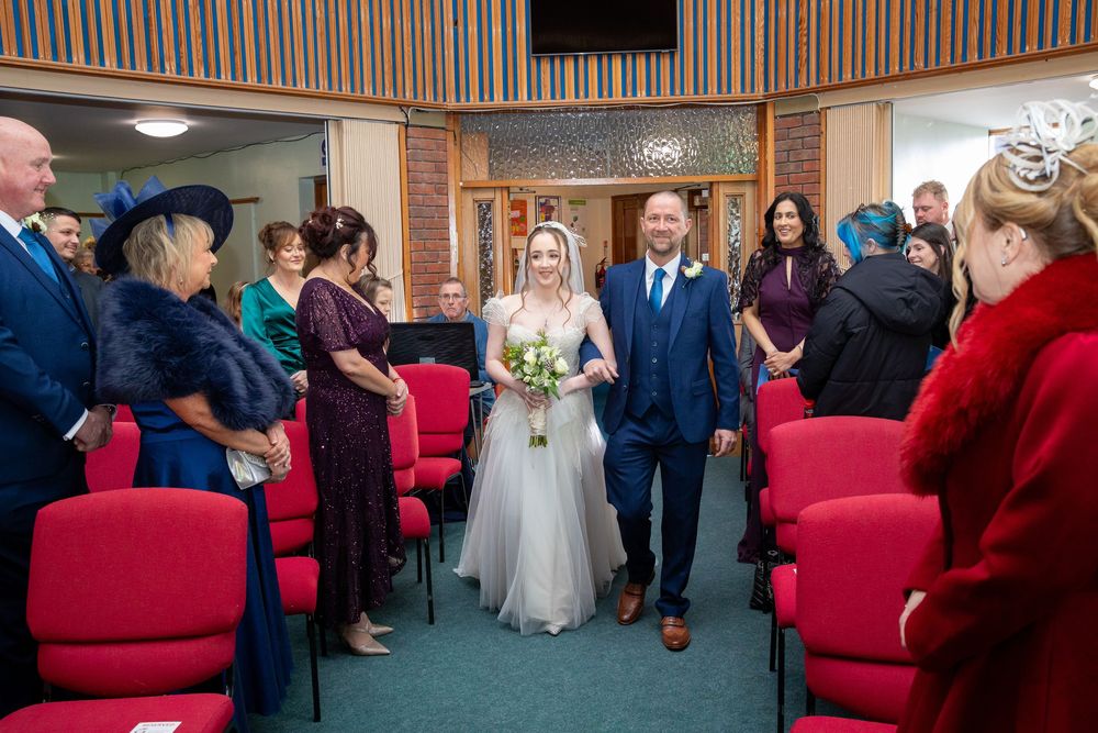 Bride and groom walk down the aisle past smiling guests in a room with red chairs.