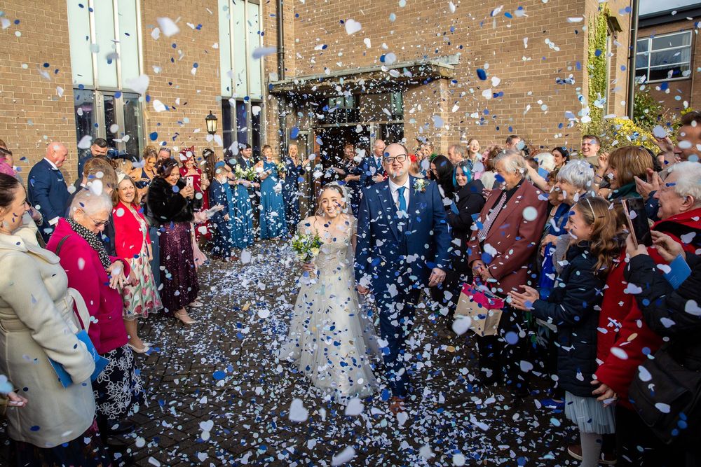 Bride and groom exit church as guests shower them with white and blue confetti outdoors.