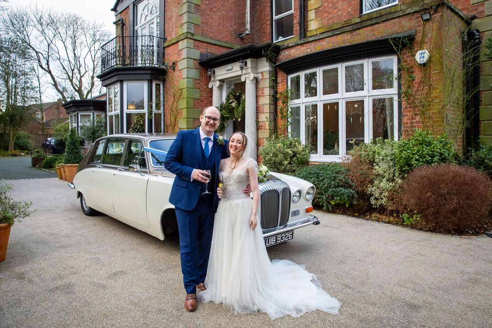 Bride and groom pose smiling beside a classic white vintage car outside an elegant manor.