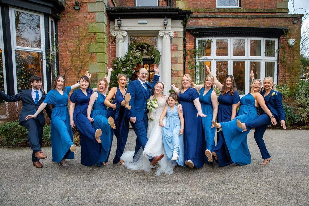 Bride groom and bridesmaids in blue dresses pose playfully outside a red brick venue.