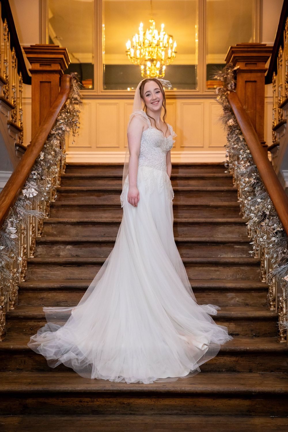 Elegant bride in lace gown poses on grand staircase with chandelier and floral decor.