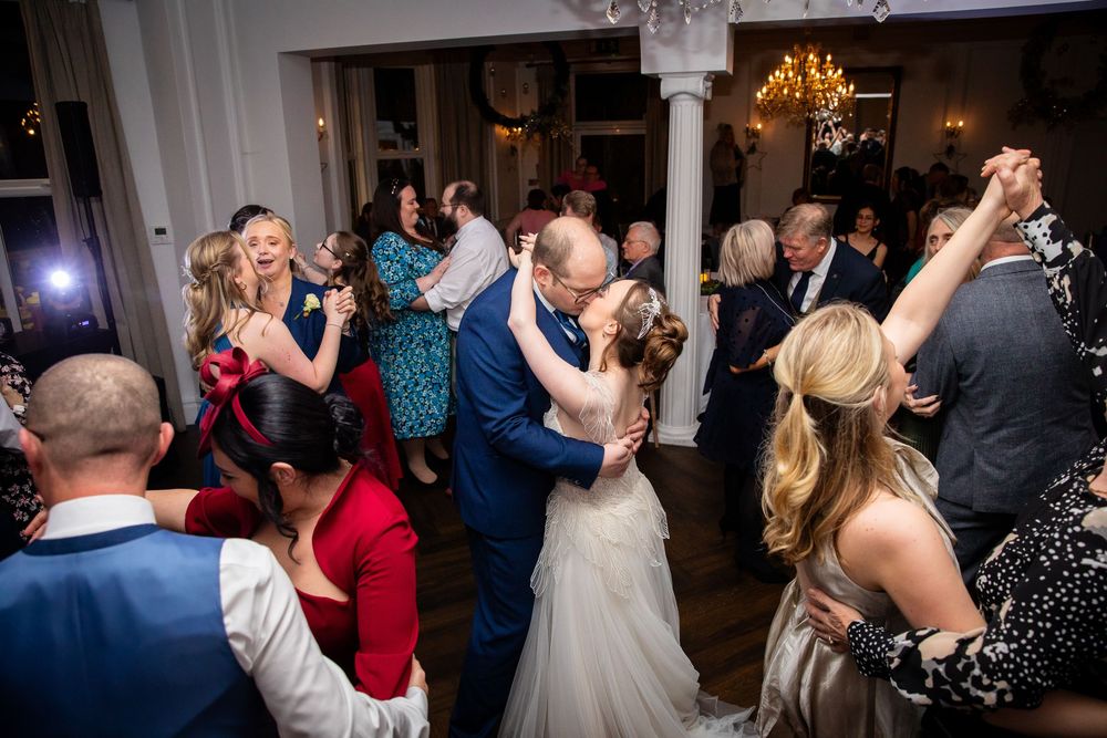 Bride and groom kiss on crowded wedding dance floor with guests dancing around them.