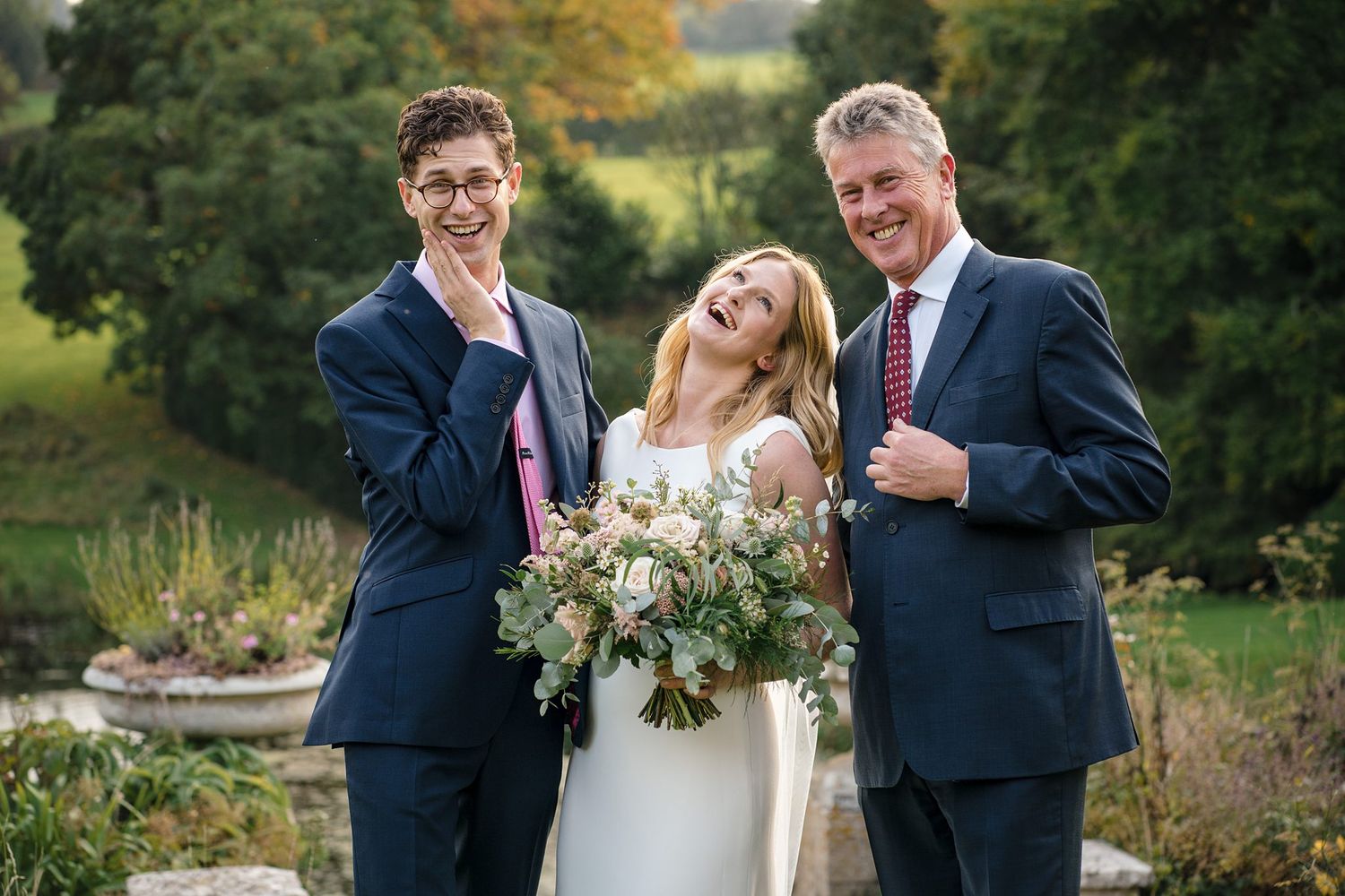 A joyful wedding party poses outdoors in navy suits and white dress with a natural garden backdrop during autumn.