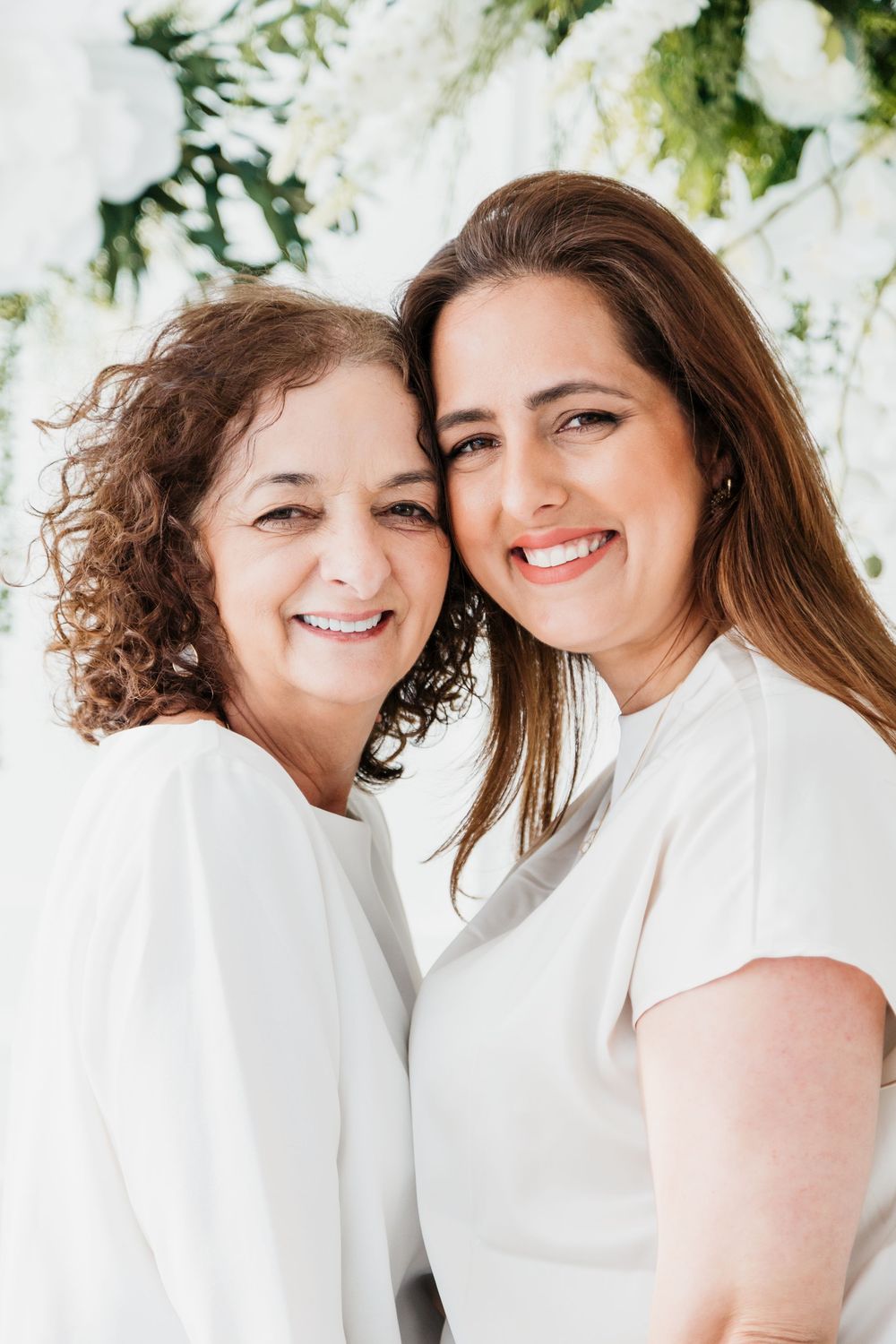 Two people in white clothing embrace and smile warmly against a bright background.
