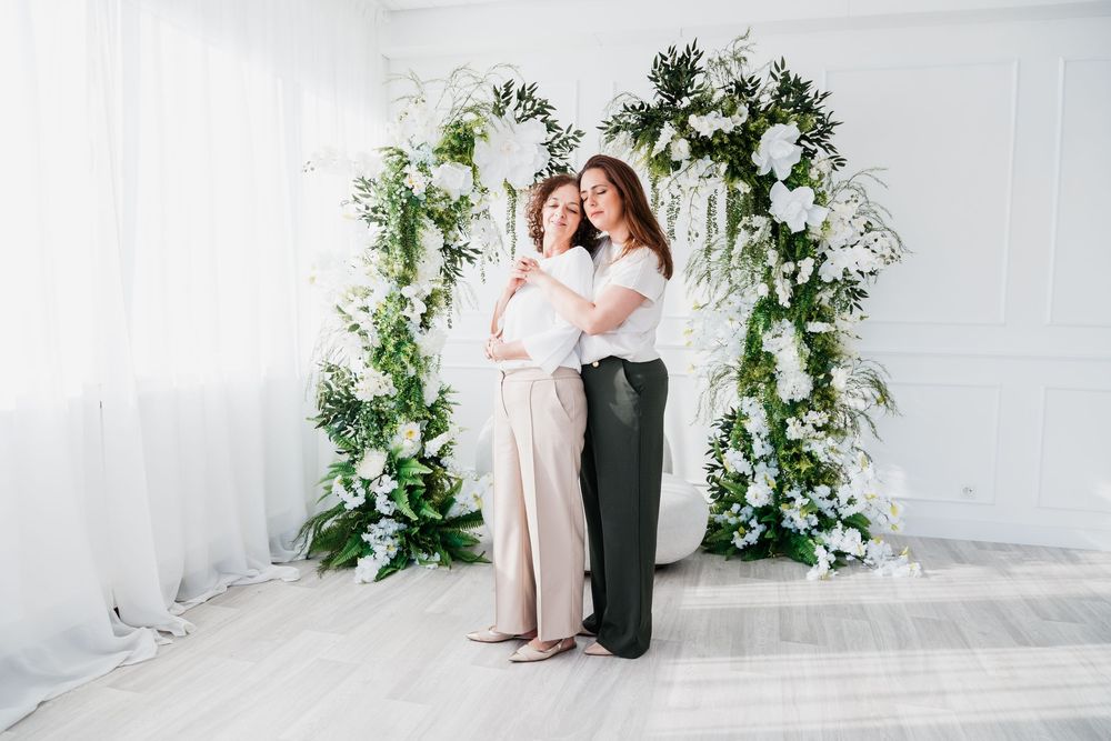 A couple poses in front of a white wall decorated with elegant green and white floral arrangements.