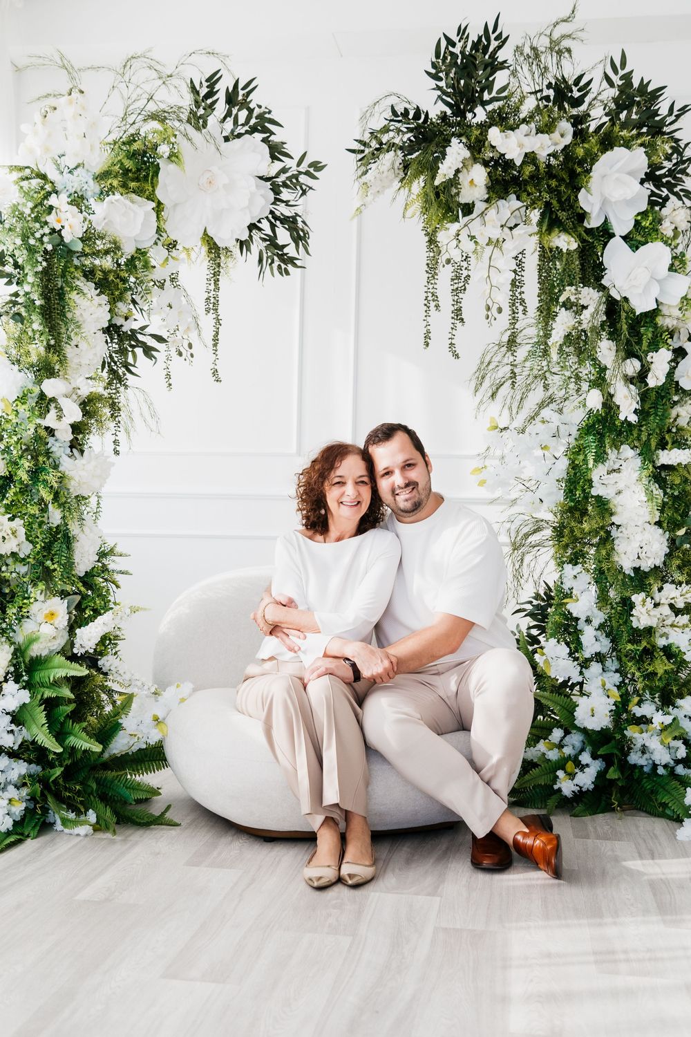 A romantic couple in white outfits poses together on a white couch surrounded by elegant floral arrangements and greenery.