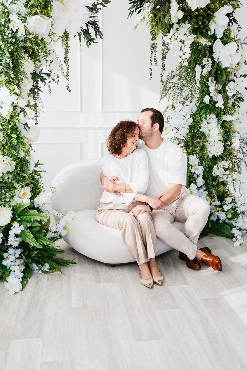 Couple in white and beige outfits share intimate moments on a white sofa surrounded by cascading greenery and flowers.