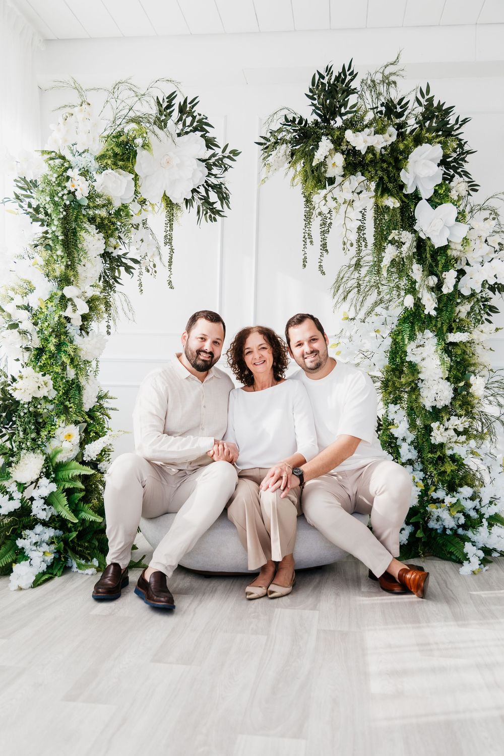 A floral arch decorated with white flowers and greenery frames three people sitting on a light-colored couch.