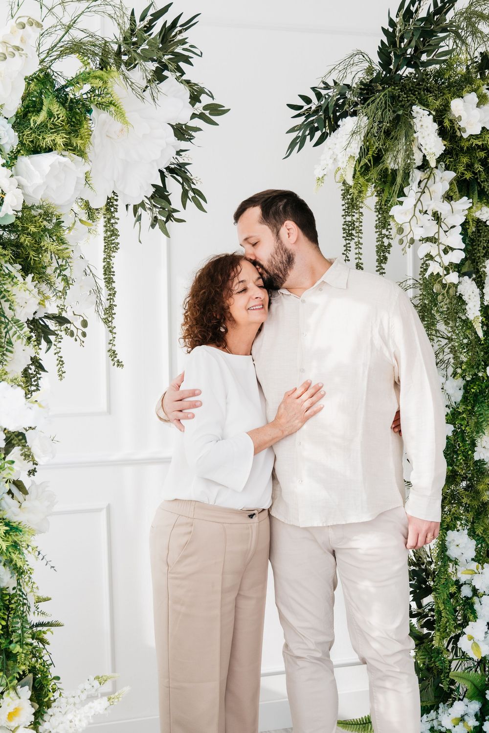 A couple embraces under a lush floral arch with white flowers and greenery against a white backdrop.
