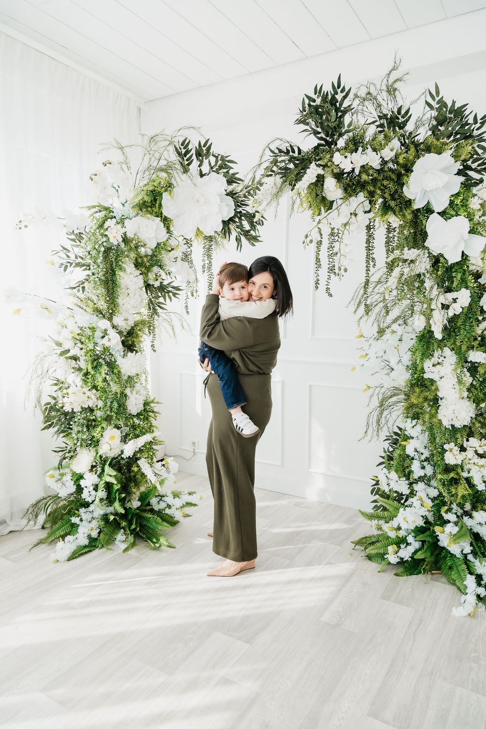 Elegant couple embraces in a bright white studio with lush greenery and floral wedding arch decorations.