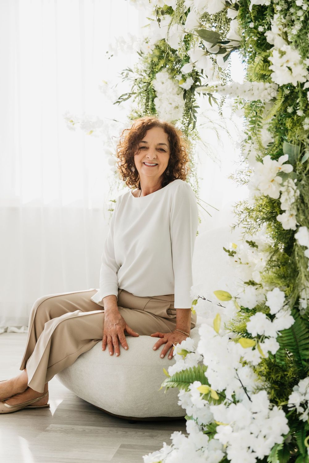 A person in white and beige clothing poses elegantly among floral decorations and greenery in a bright studio setting.