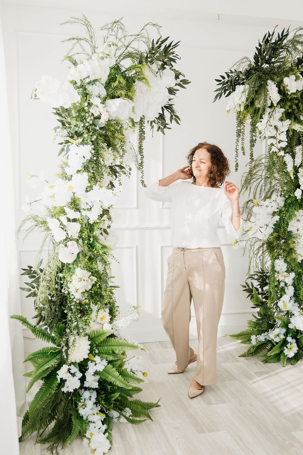 Elegant white and green floral arch installation with ferns and flowers decorates a bright interior space with light wood floors.
