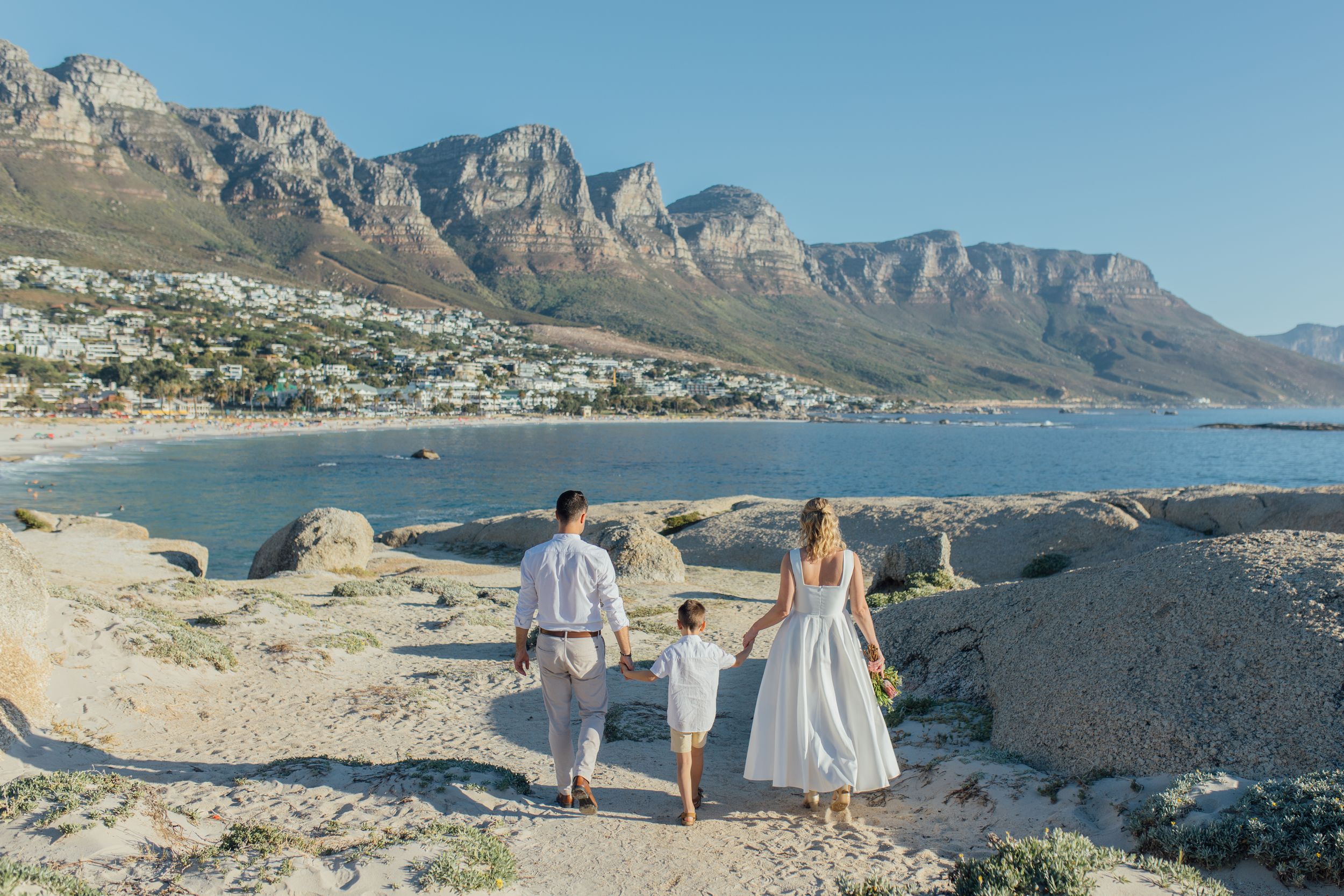 Family walking hand-in-hand along rocky Camps Bay beach with Twelve Apostles mountain range and Atlantic Ocean in background.