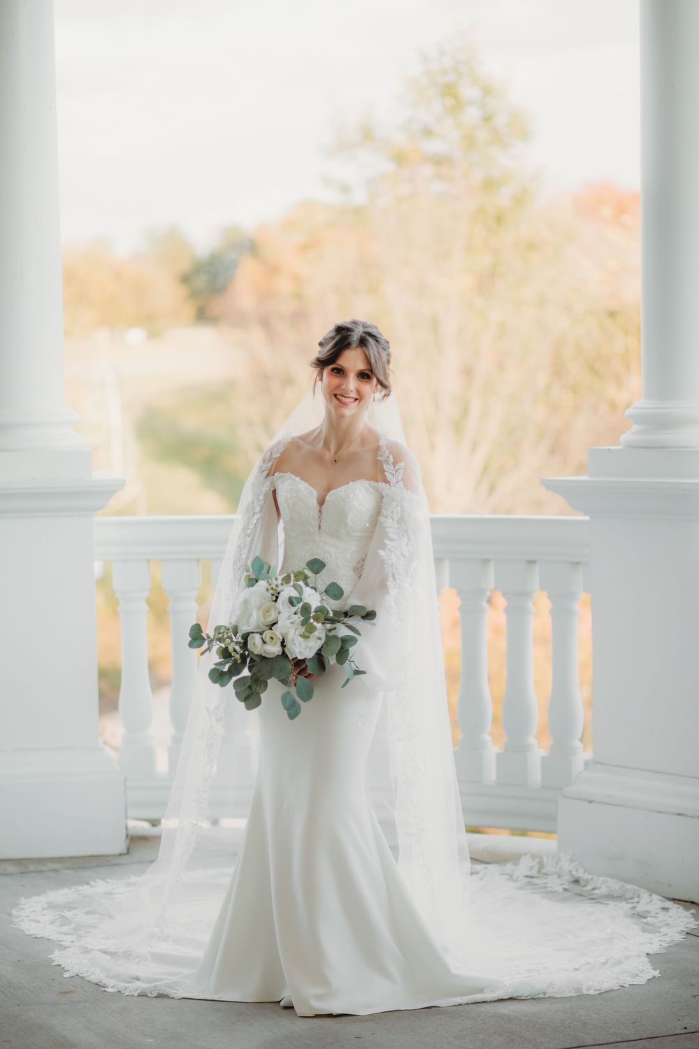 Bride in elegant white wedding gown with long train and veil poses on classic white columned porch in natural light in huntsville alabama