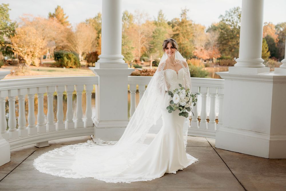 A bride in a flowing white gown with long train holds a bouquet while standing on an elegant white columned porch.