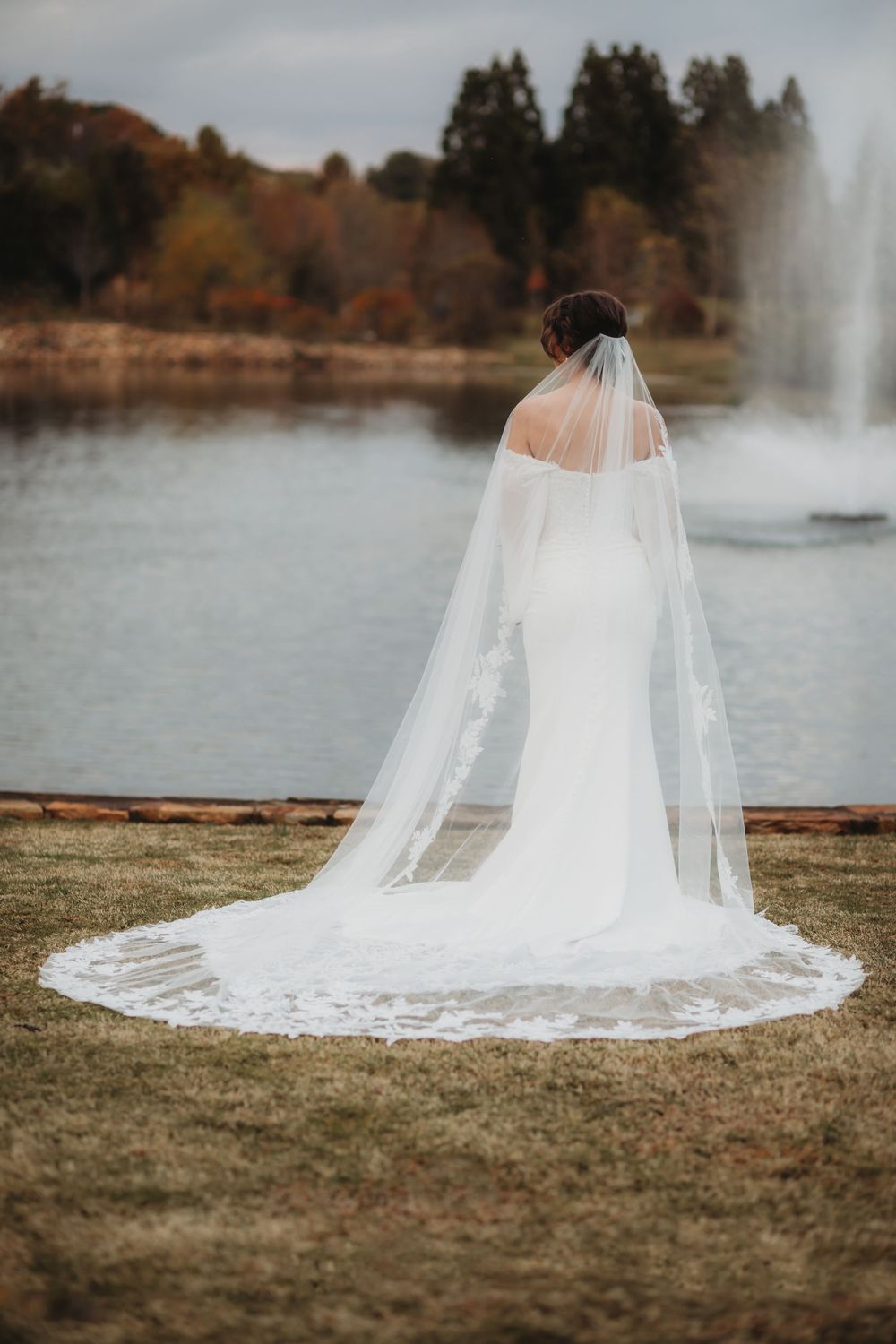 A flowing cathedral-length bridal veil with lace trim spreads elegantly across grass near a lake with fountains in Huntsville, Alabama