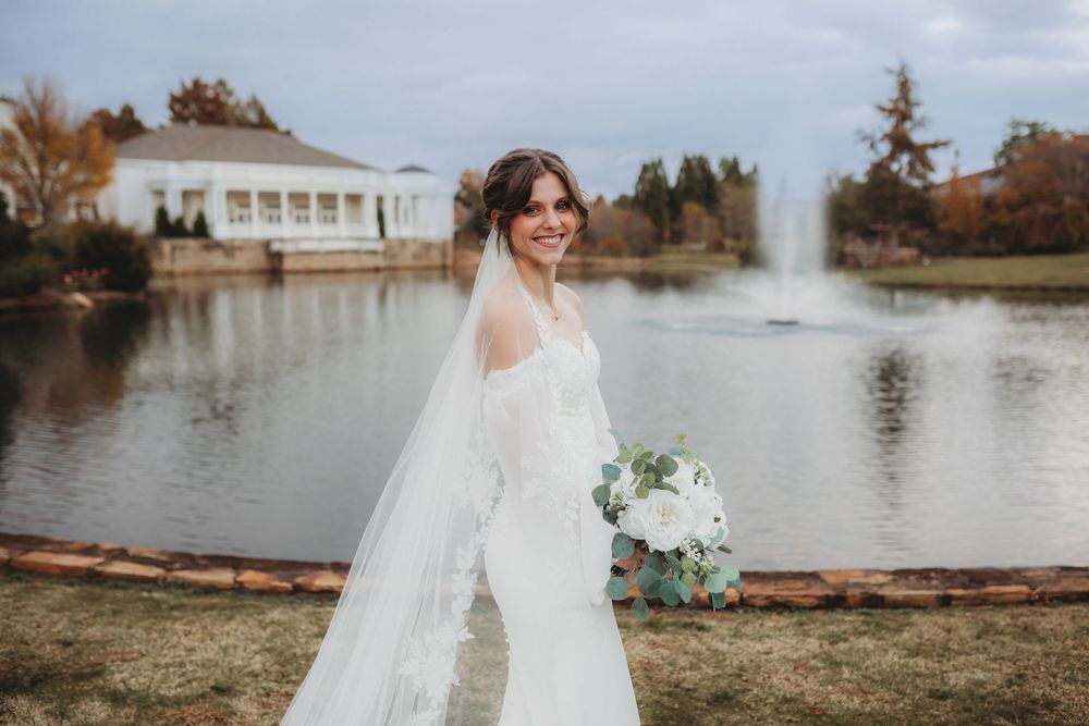 Bride in a flowing white wedding gown stands by a scenic lake with autumn trees and a fountain in the background in Huntsville Alabama.