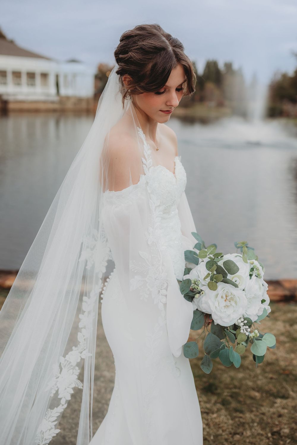 A bride in a white lace wedding dress and veil holds a bouquet of white flowers and eucalyptus by a lake in Huntsville Alabama.