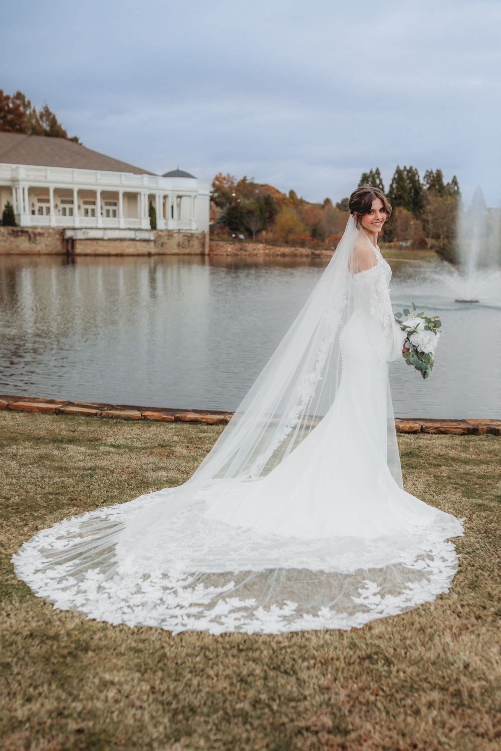 A stunning cathedral length lace veil trails behind a wedding dress on grass near a waterfront venue in Huntsville Alabama.