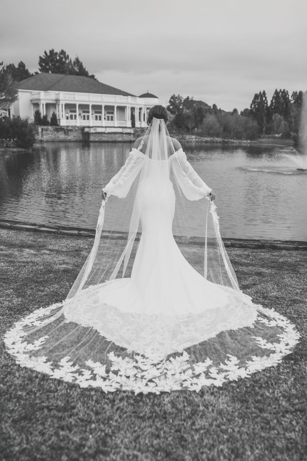 A white wedding dress with a long train and veil laid out on grass by a lakeside building in black and white in Huntsville Alabama
