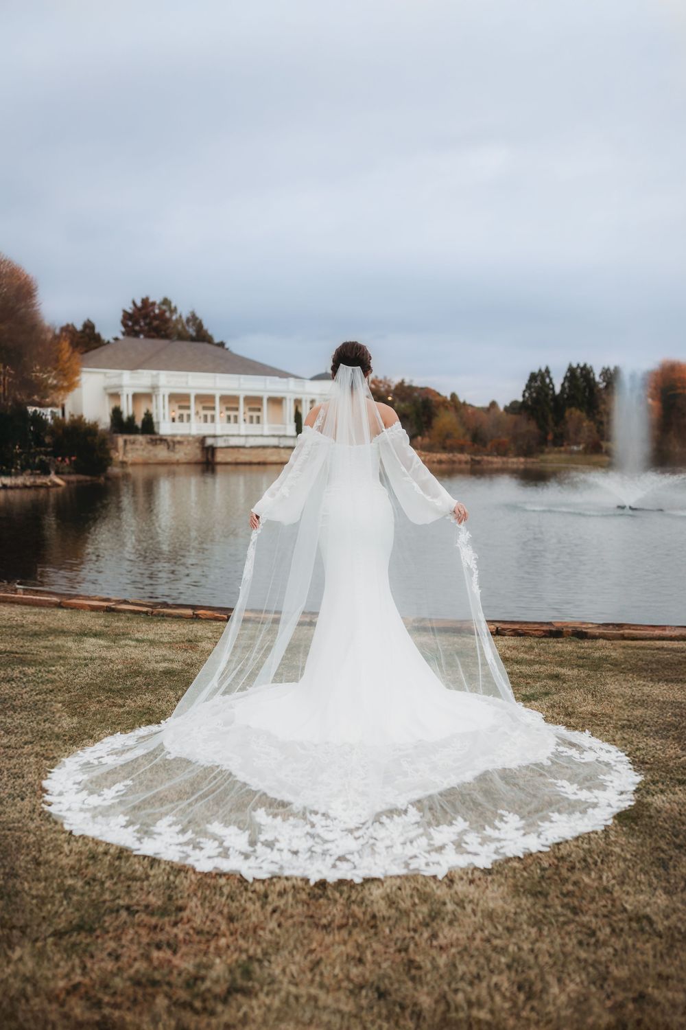 A stunning cathedral-length lace wedding veil spreads across green grass by a serene lakeside setting in Hunstville Alabama.