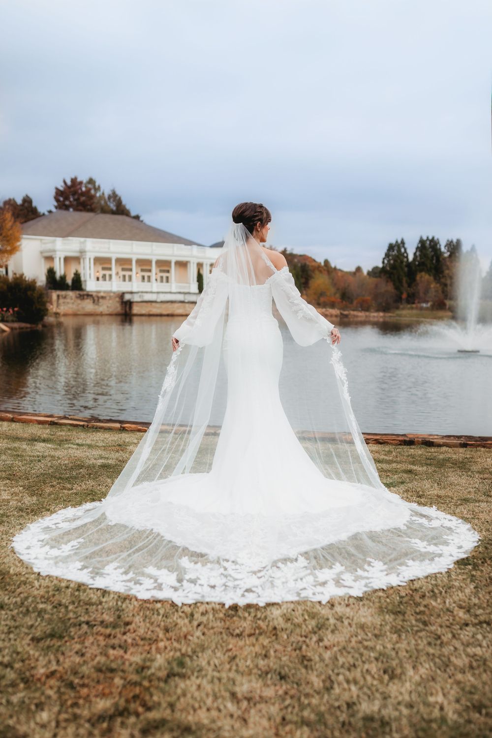 A flowing white cathedral-length wedding veil with lace details spreads across grass near a lakeside venue in Huntsville Alabama