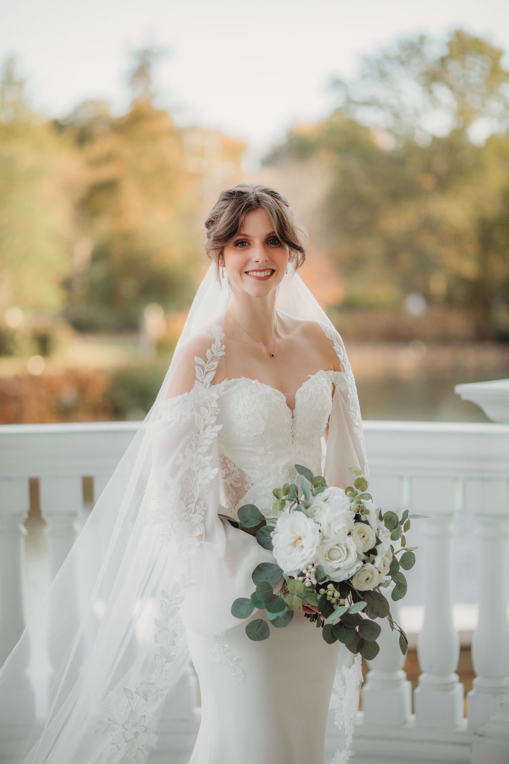 Elegant bride in lace veil and white gown holds white floral bouquet with eucalyptus on outdoor balcony in autumn in Huntsville alabama.