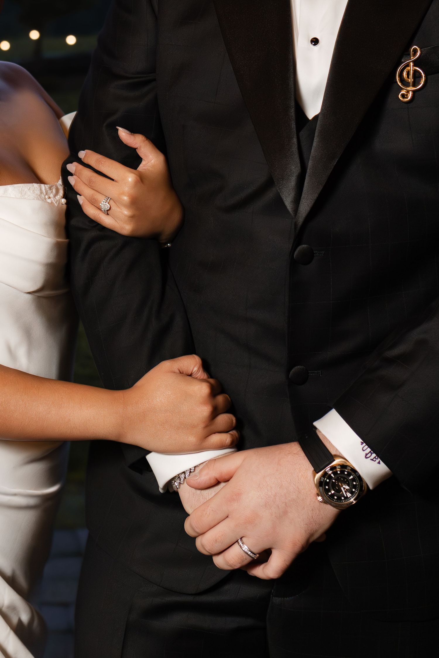 Close-up detail of formal wedding attire showing black tuxedo, white cuffs, wedding rings and a wristwatch.