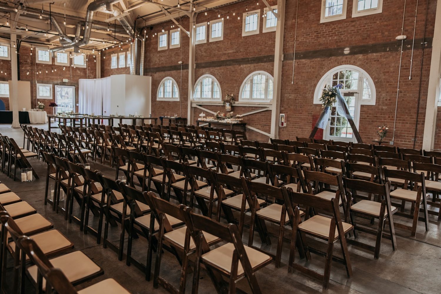Rows of dark wooden folding chairs arranged in a historic brick building with arched windows and industrial details.