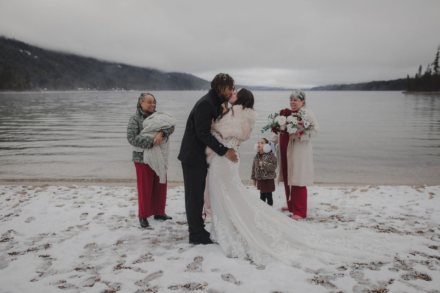 A winter wedding ceremony takes place on a snowy beach beside a serene lake with mountains in the background.