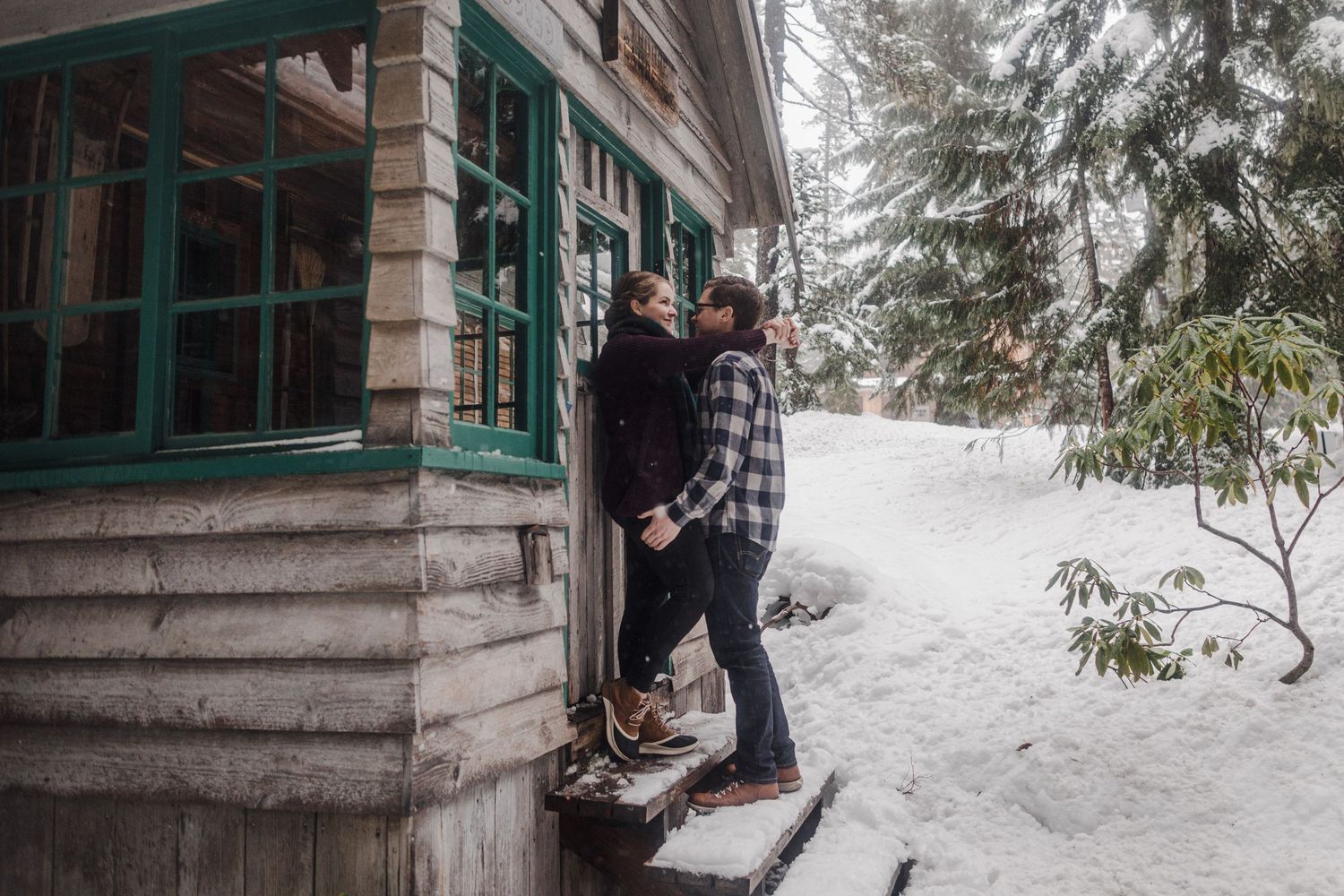 A couple shares a romantic moment on the steps of a rustic cabin surrounded by snowy winter scenery.