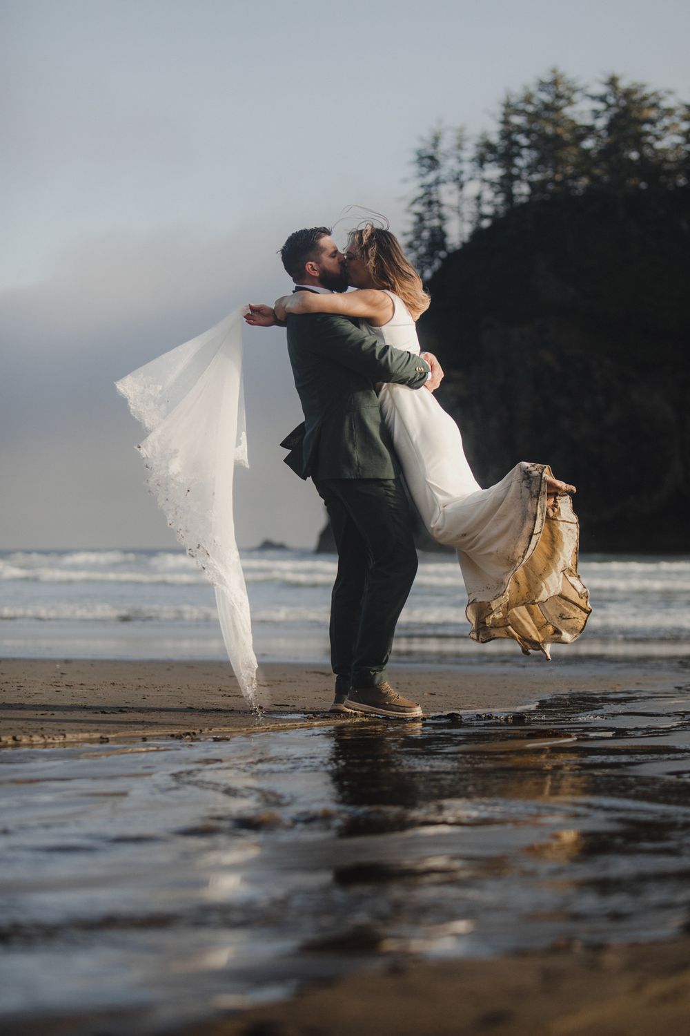 A couple in wedding attire shares a romantic dip kiss on a misty beach at sunset with waves in the background.